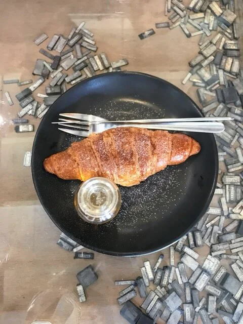 Croissant dusted with icing sugar on a black plate, with a fork and a small glass jar of jam.