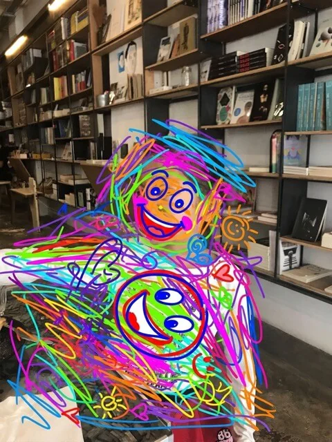 Someone standing in front of the bookshelves holding up a drawing, both covered over with a scribble of coloured pen and two smiling faces.