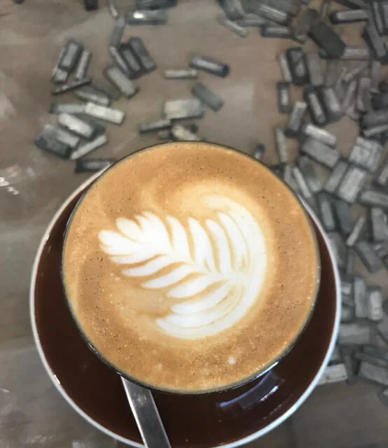 Flat white with a rosetta poured on top, in a dark brown cup and saucer, on a table of loose lead type.