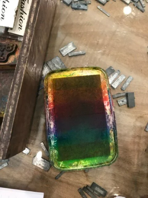Stamp pad inked in bands of green, blue, red and yellow, on a wooden table scattered with lead type.