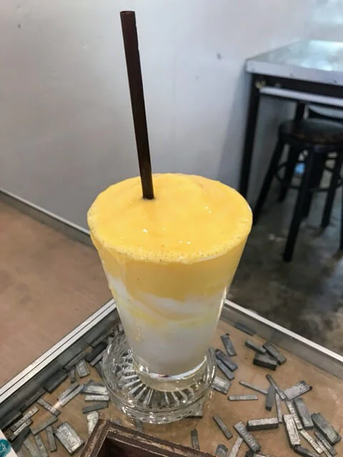 Tall glass of mango smoothie layered over yoghurt with a wooden straw, on a table of loose lead type.