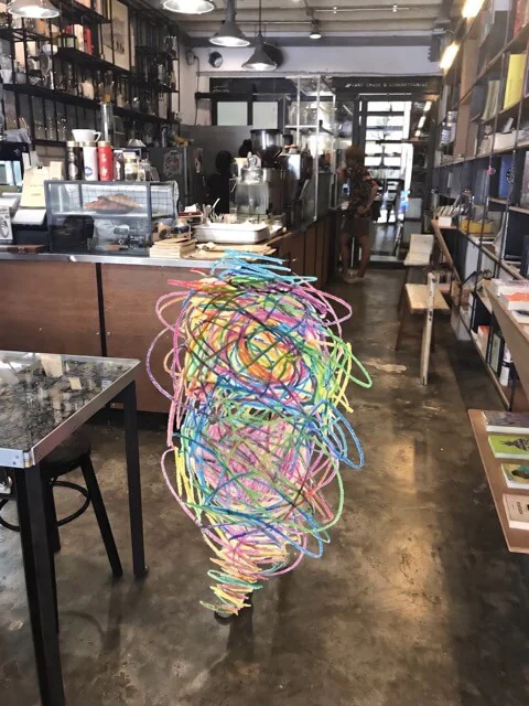 The shop looking back from the door: coffee counter on the left, shelves down the right-hand wall, and the room running back to a doorway. A figure in the middle of the frame is scribbled over in colour.