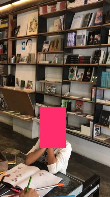 A child reading at a glass-topped table below shelves of books stacked to the ceiling, an open sketchbook and coloured pencils in front of them. Their face is covered by a pink block.