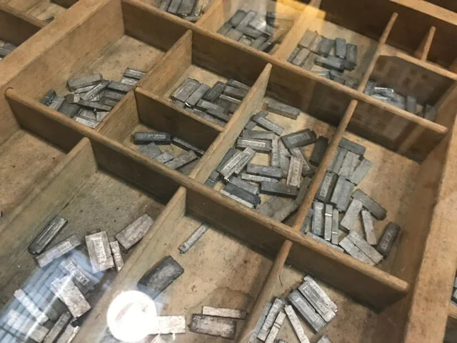 Lead type sorted into the compartments of a wooden printer's tray, under the glass of a café table.